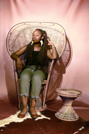 A photo of a Black teenage girl wearing a black shirt, green pants, and brown shoes sitting in a wicker chair in front of a draped pink cloth. Her hair is braided, and she is wearing a headband, Africa earrings, a necklace, and an anklet. Next to the wicker chair is a small wicker table. The chair and table are on a brown and white hide. She has one hand draped over the chair and one placed at her head. Her head is tilted upwards and to the side with her eyes looking forward.